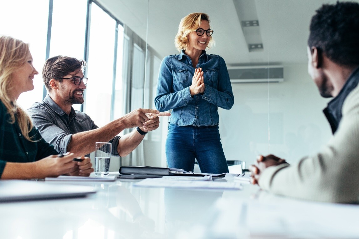 Frau steht lächelnd vor Gruppe an Konferenztisch.
