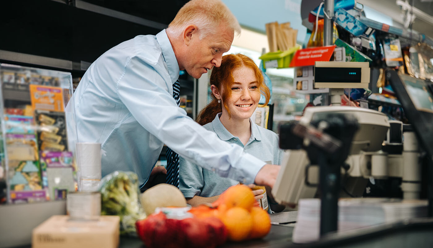 Junge Frau an der Kasse im Supermarkt, die von einem Kollegen eingewiesen wird